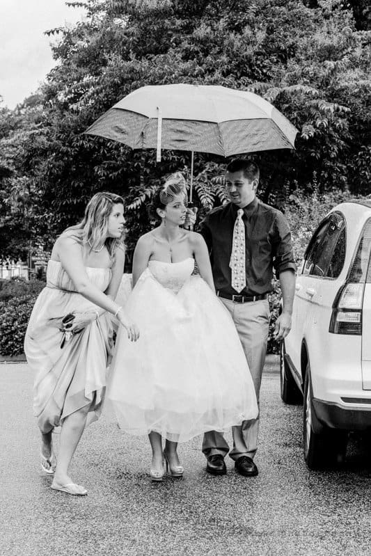 A black-and-white photo of a bride in a wedding dress being assisted by two people as she walks. One person holds an umbrella over her head. Theyre outside near a car, with trees in the background.