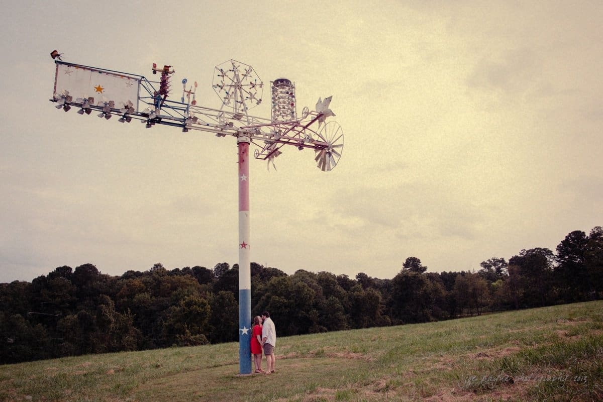 A couple stands at the base of a large, colorful wind-powered sculpture on a grassy hillside. The sculpture features various geometric shapes and elements. Trees and a cloudy sky form the background.