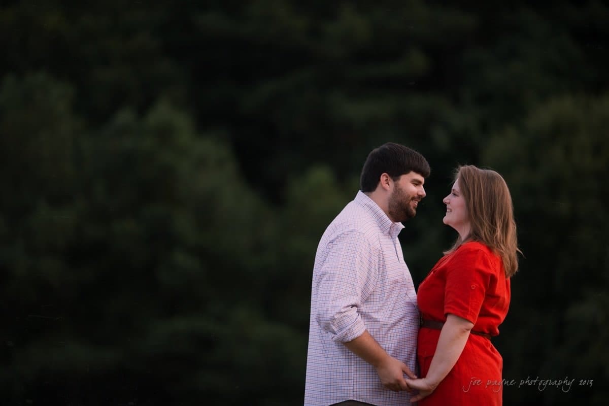 A couple holds hands and smiles at each other in a lush green outdoor setting. The man wears a light shirt, and the woman is in a red dress. The background is blurred with trees, creating a romantic atmosphere.