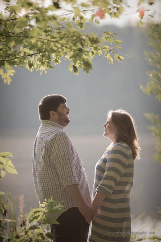 A couple holds hands and smiles at each other in a sunlit outdoor setting. The man wears a plaid shirt, and the woman wears a striped dress. They are surrounded by greenery, with soft, natural light creating a warm atmosphere.