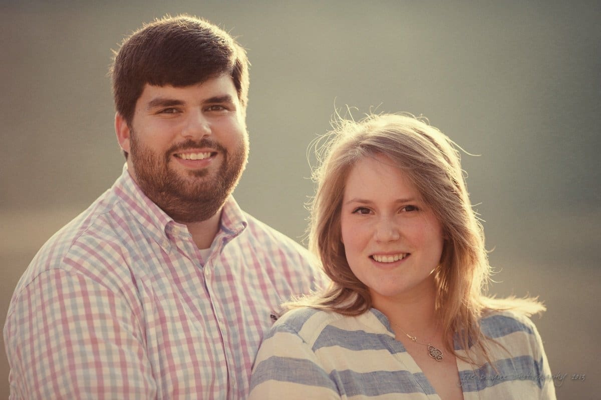 A smiling man and woman stand side by side outdoors, bathed in warm, soft lighting. The man wears a plaid shirt, while the woman has a striped shirt with her hair gently tousled by the wind. The background is blurred and natural.