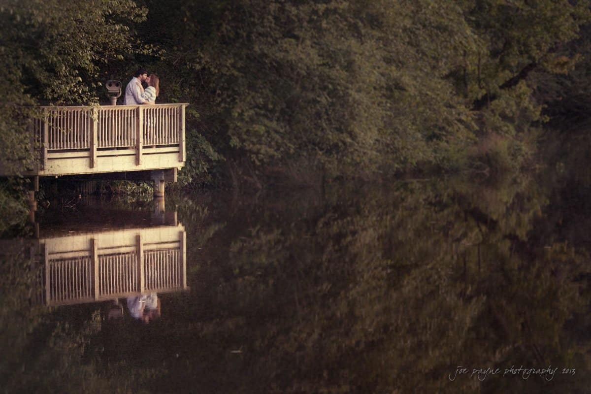 A couple stands embracing on a wooden deck overlooking a calm, reflective lake surrounded by lush greenery. The scene is peaceful and serene, capturing a moment of intimacy and tranquility in nature.