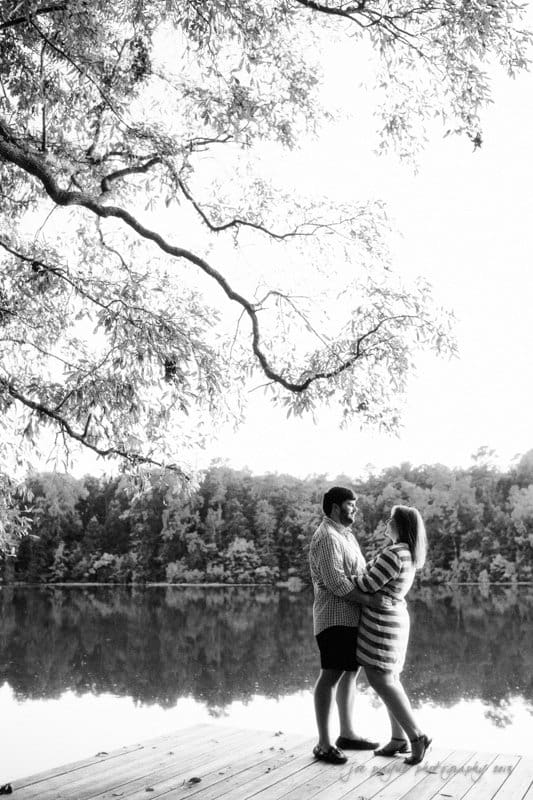 A black and white photo of a couple standing on a wooden dock by a calm lake, surrounded by trees. They are facing each other, holding hands and smiling, with branches arching above them.
