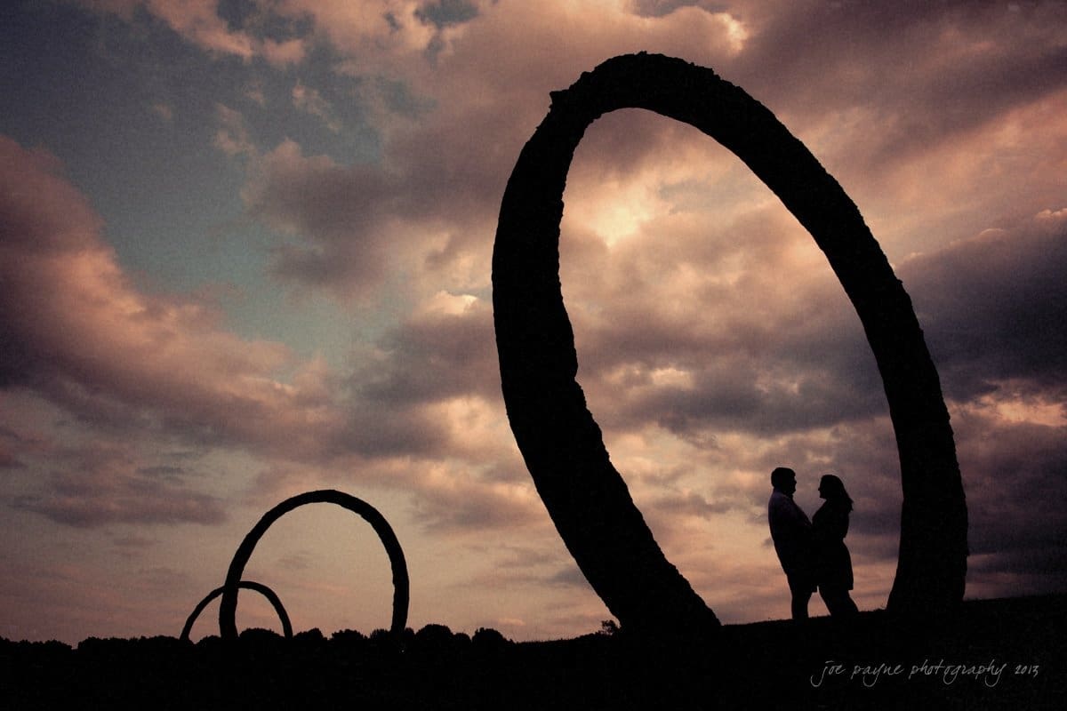 Silhouetted couple stands near large circular sculptures at sunset. The sky is filled with dramatic clouds, creating a serene and romantic scene.