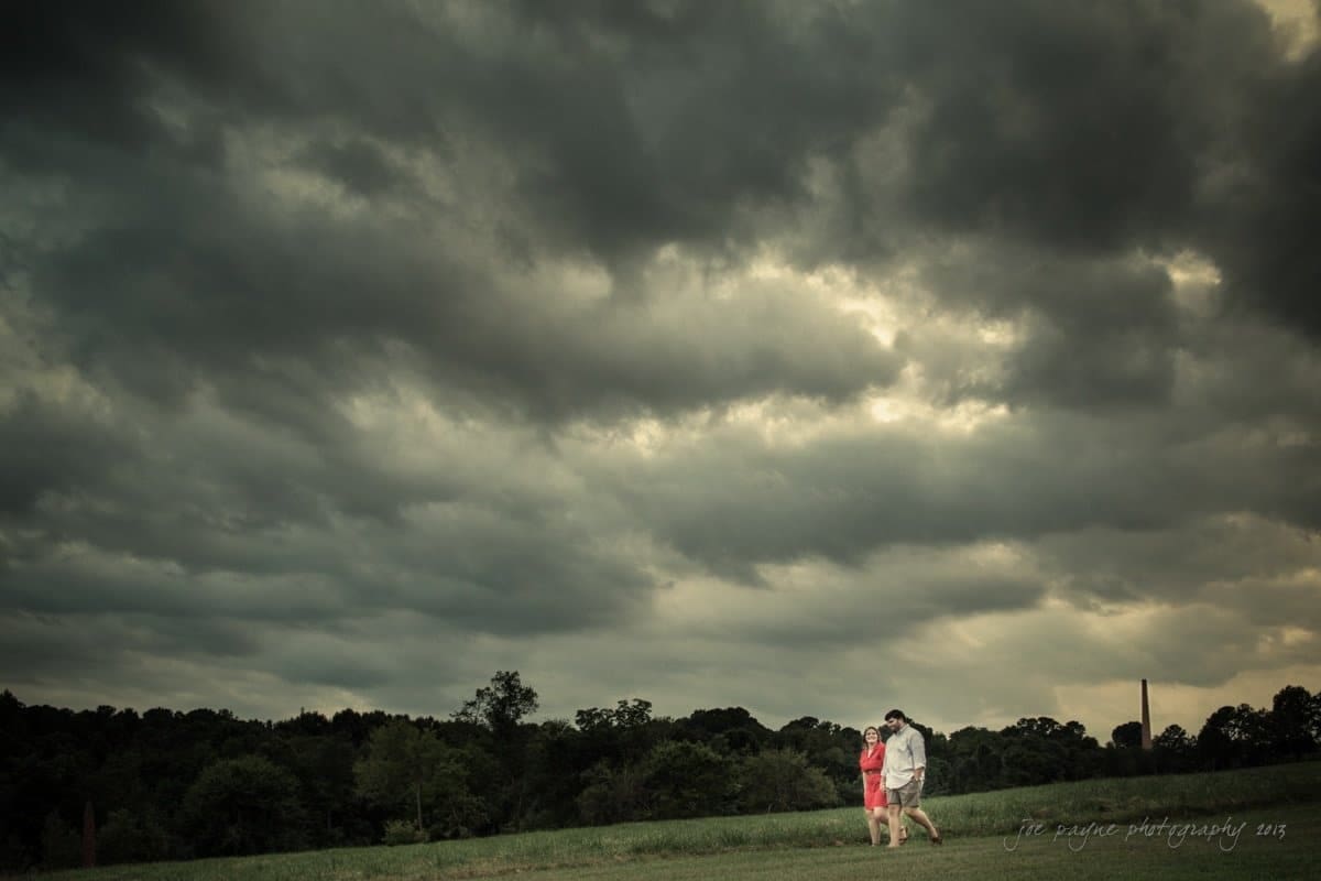 A couple walks hand in hand across a grassy field under a dramatic, cloudy sky. Dark, thick clouds create a moody atmosphere, contrasting with the green landscape and the couples bright clothing.