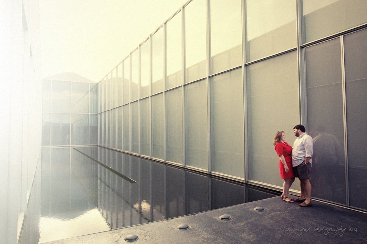 A couple stands at the edge of a reflective pool, surrounded by tall, modern, gray walls. The woman wears a red dress, and the man is dressed in a white shirt and shorts. The scene is serene and minimalist.