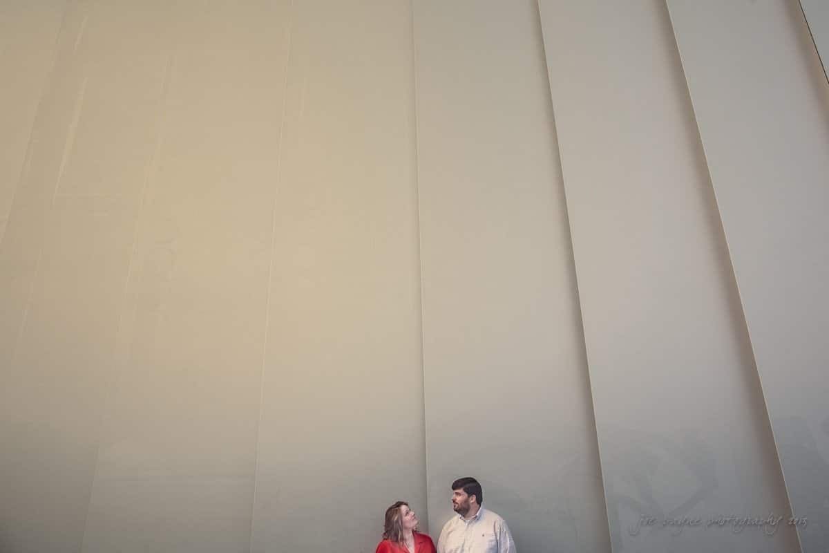 A man and a woman stand facing each other at the base of a large, textured wall with a series of vertical panels, giving a sense of scale and depth. The wall is light beige, and the space is open and minimalist.