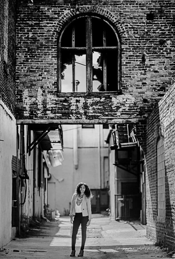 A person with curly hair stands in an alleyway beneath a large, arched brick window. The alley is flanked by brick and concrete walls, and sunlight filters through the open space above. The scene is in black and white.