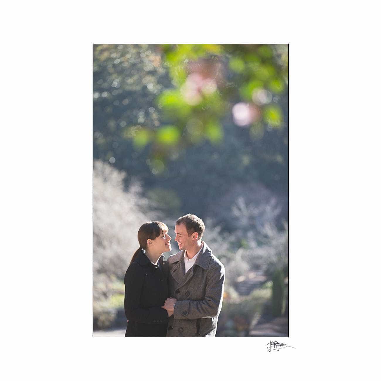 duke chapel engagement photography ~ brianna and chris A couple stands together in an outdoor setting, looking at each other and smiling. They are dressed in coats, and the background is softly blurred with greenery and light.