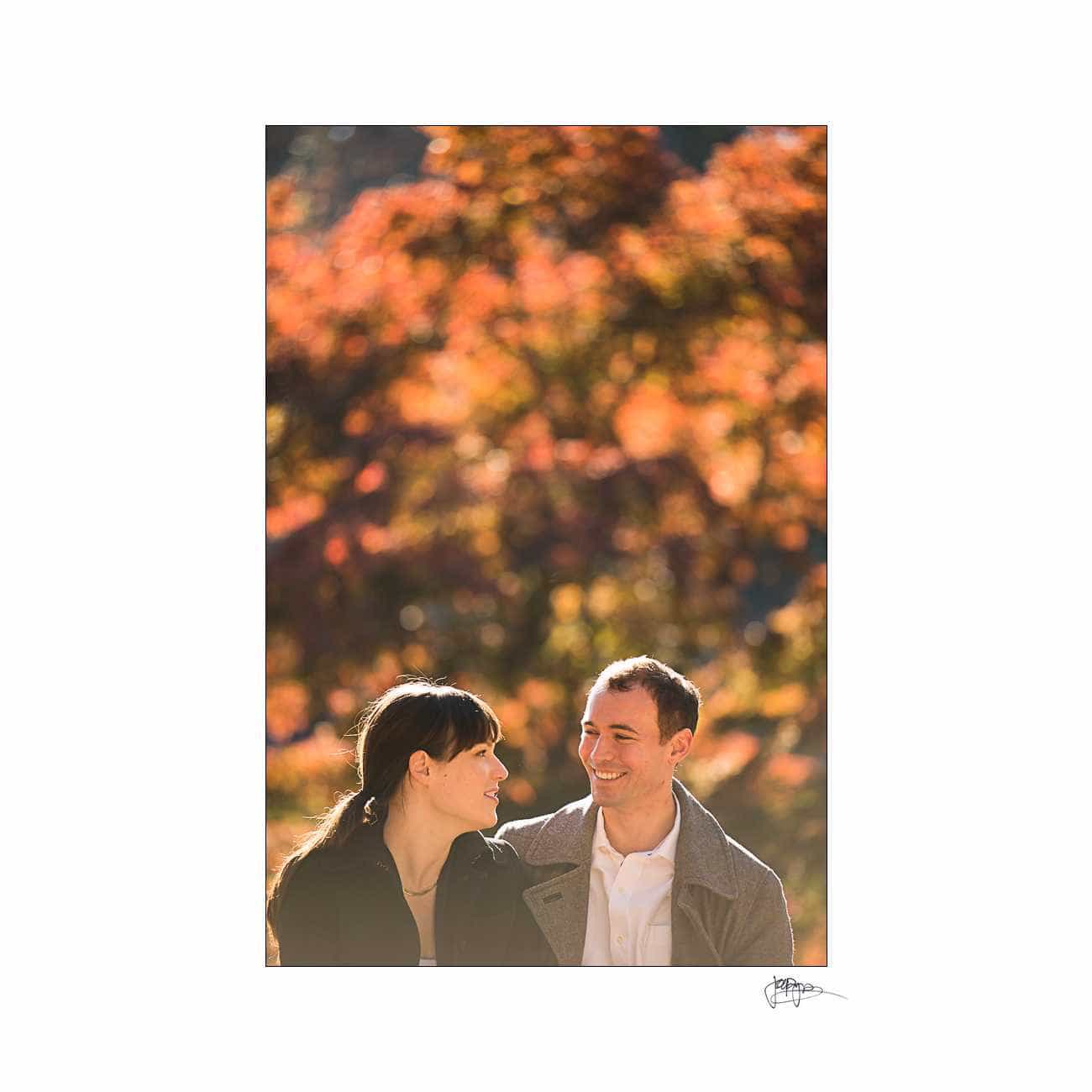 duke chapel engagement photography ~ brianna and chris A couple stands close together, smiling, with a backdrop of vibrant autumn leaves in shades of orange and red. The sunlight casts a warm glow over the scene.