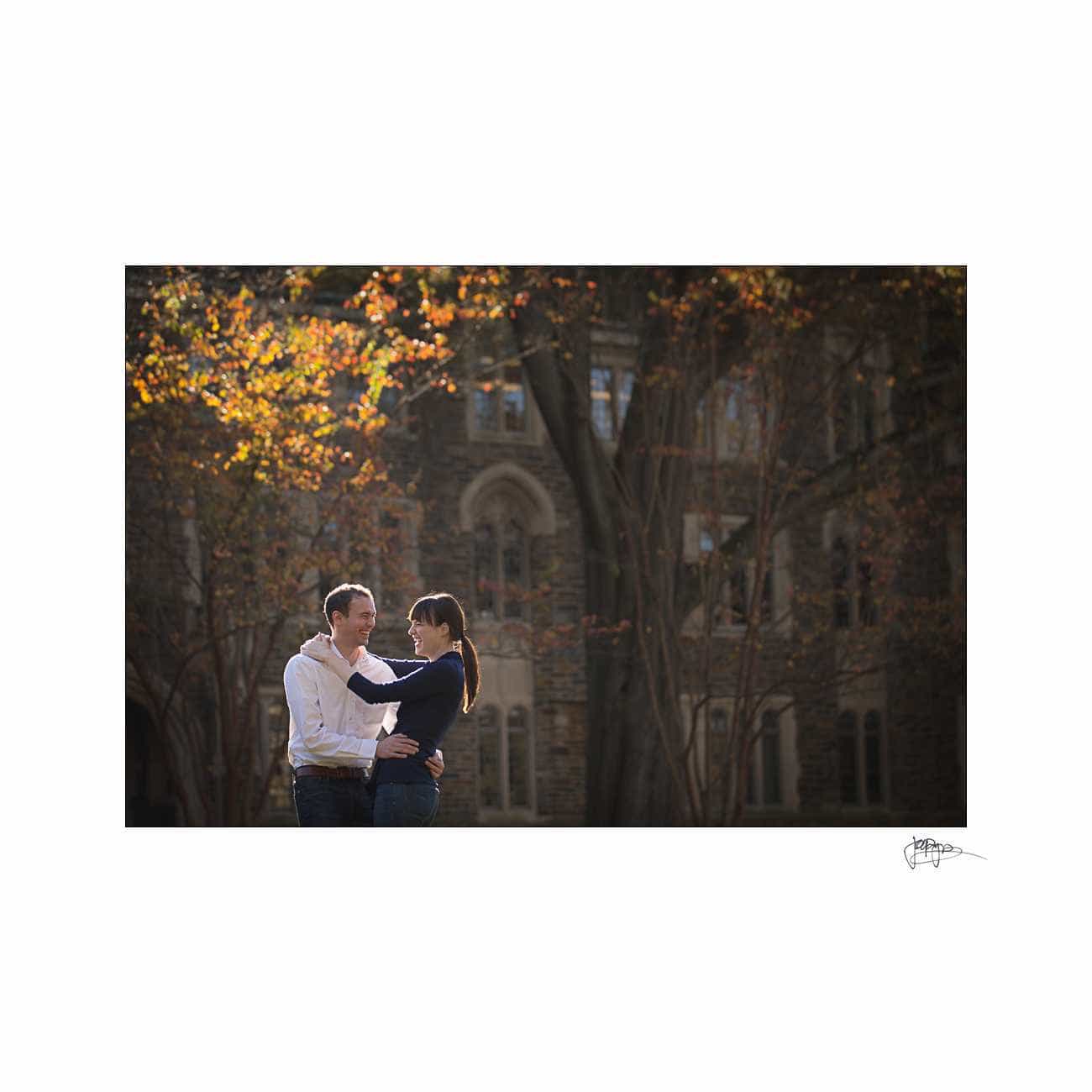 A couple stands embracing in a sunlit courtyard surrounded by trees with autumn leaves. A historic stone building is in the background, creating a serene and romantic atmosphere.