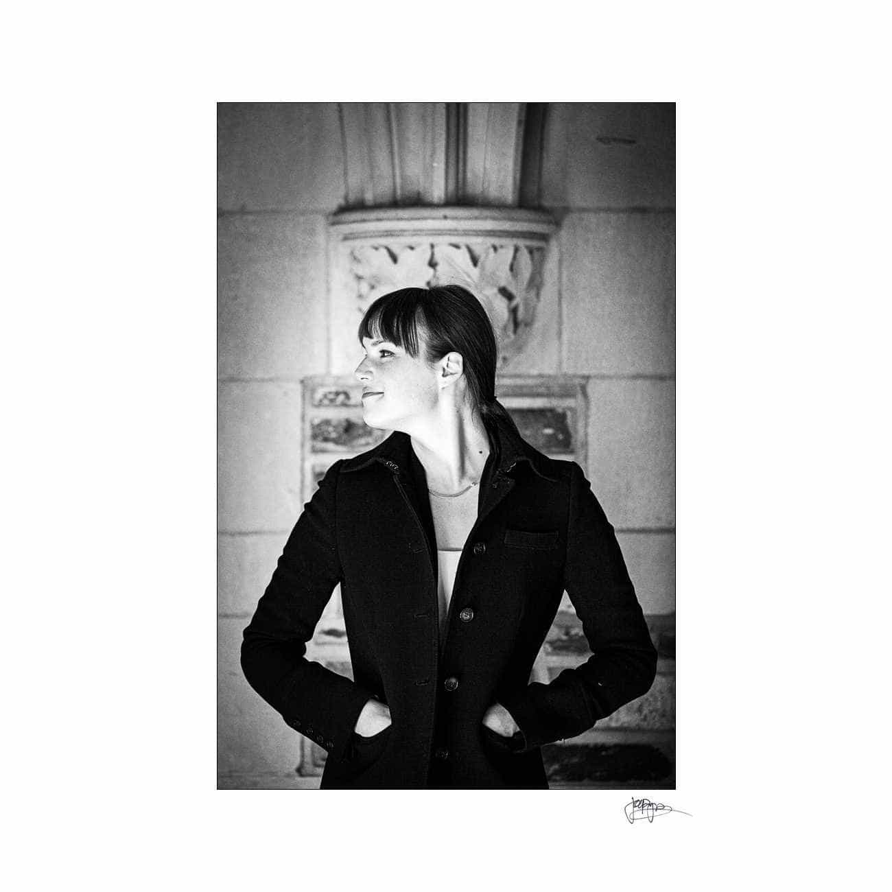 duke chapel engagement photography ~ brianna and chris A woman in a black coat stands against a decorated stone wall, looking to the side with her hands in her pockets. The image is in black and white, capturing a timeless, contemplative moment.