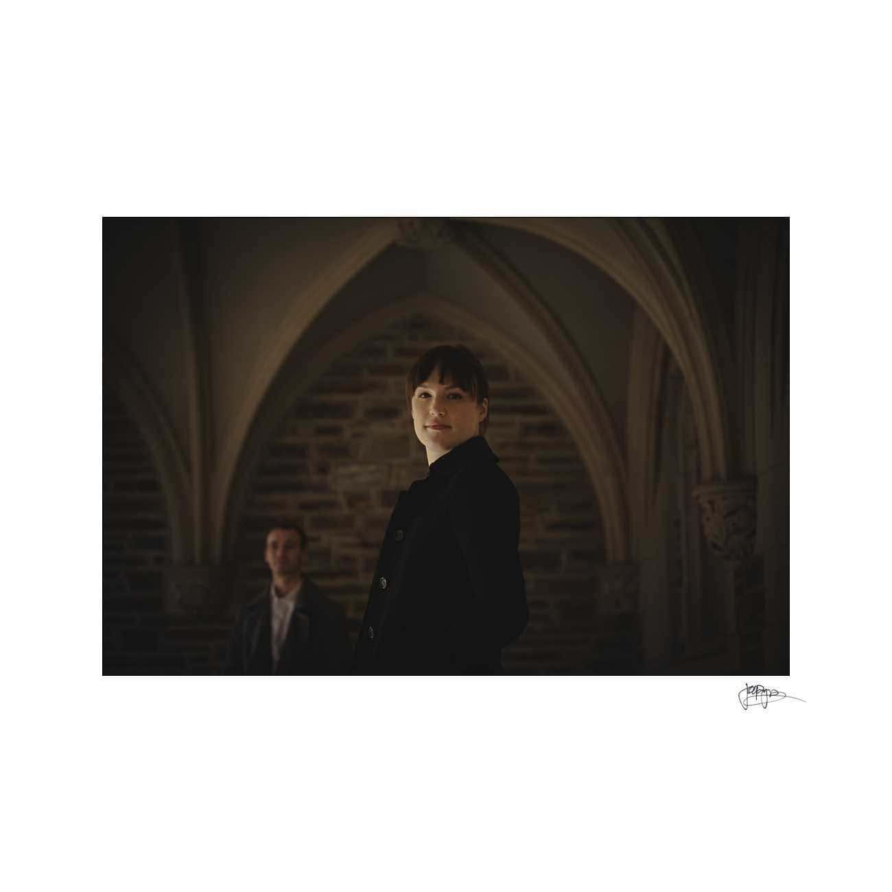 duke chapel engagement photography ~ brianna and chris A woman in a dark coat stands under an arched, dimly lit stone ceiling, gazing forward with a slight smile. In the background, a man in a suit stands out of focus, adding depth to the scene.
