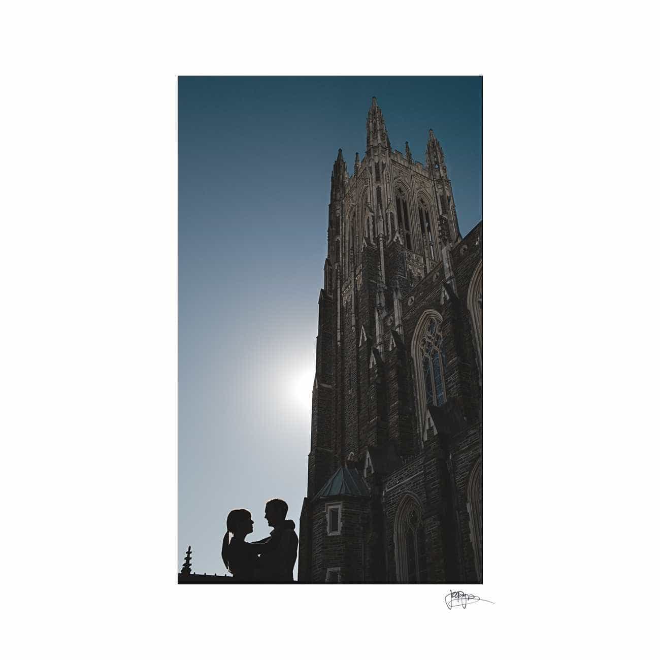 duke chapel engagement photography ~ brianna and chris Silhouette of a couple embracing in front of a tall, gothic-style cathedral with a pointed spire against a bright sky. The sun is partially obscured, creating a soft halo effect around the architecture.