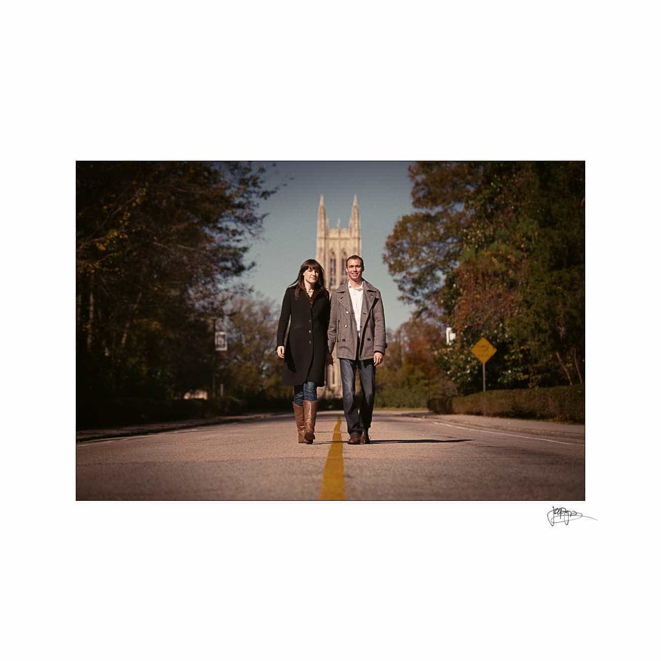 duke chapel engagement photography ~ brianna and chris A man and woman walk side by side down a tree-lined road. They are dressed in fall attire. In the background, a tall, Gothic-style building is visible. The road has a yellow dividing line, and the scene is set in a clear, sunny day.