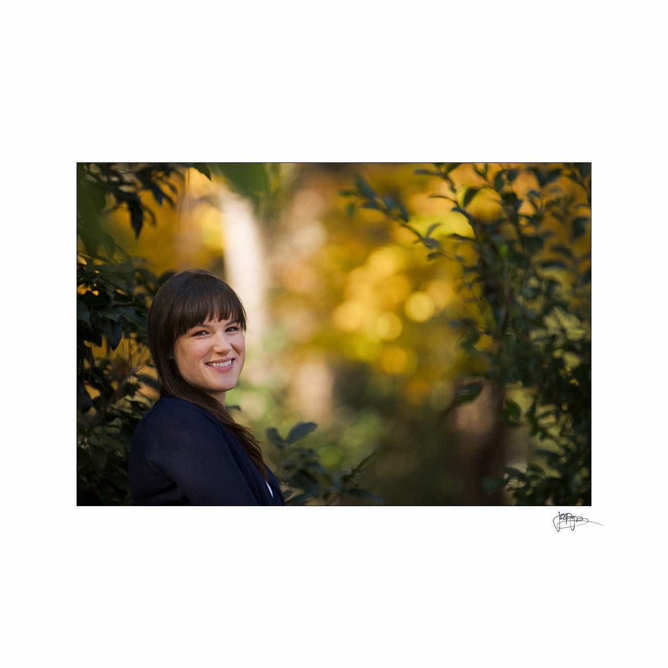 duke chapel engagement photography ~ brianna and chris A woman with long brown hair smiles while standing amidst green foliage. The background features blurred yellow and green leaves, suggesting a natural, outdoor setting.
