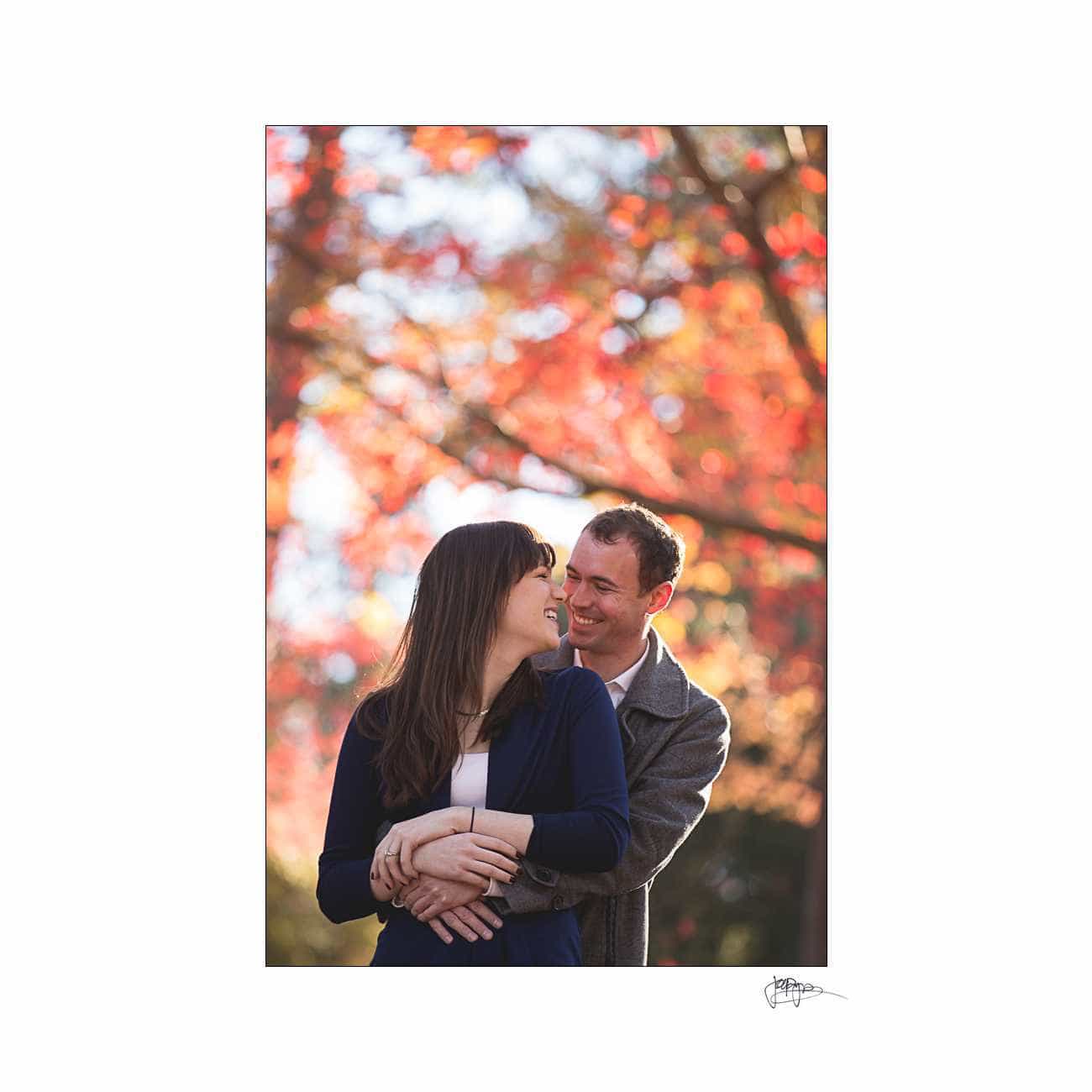 duke chapel engagement photography ~ brianna and chris A couple stands in a park with vibrant autumn leaves in the background. The man embraces the woman from behind, and they both smile warmly at each other.