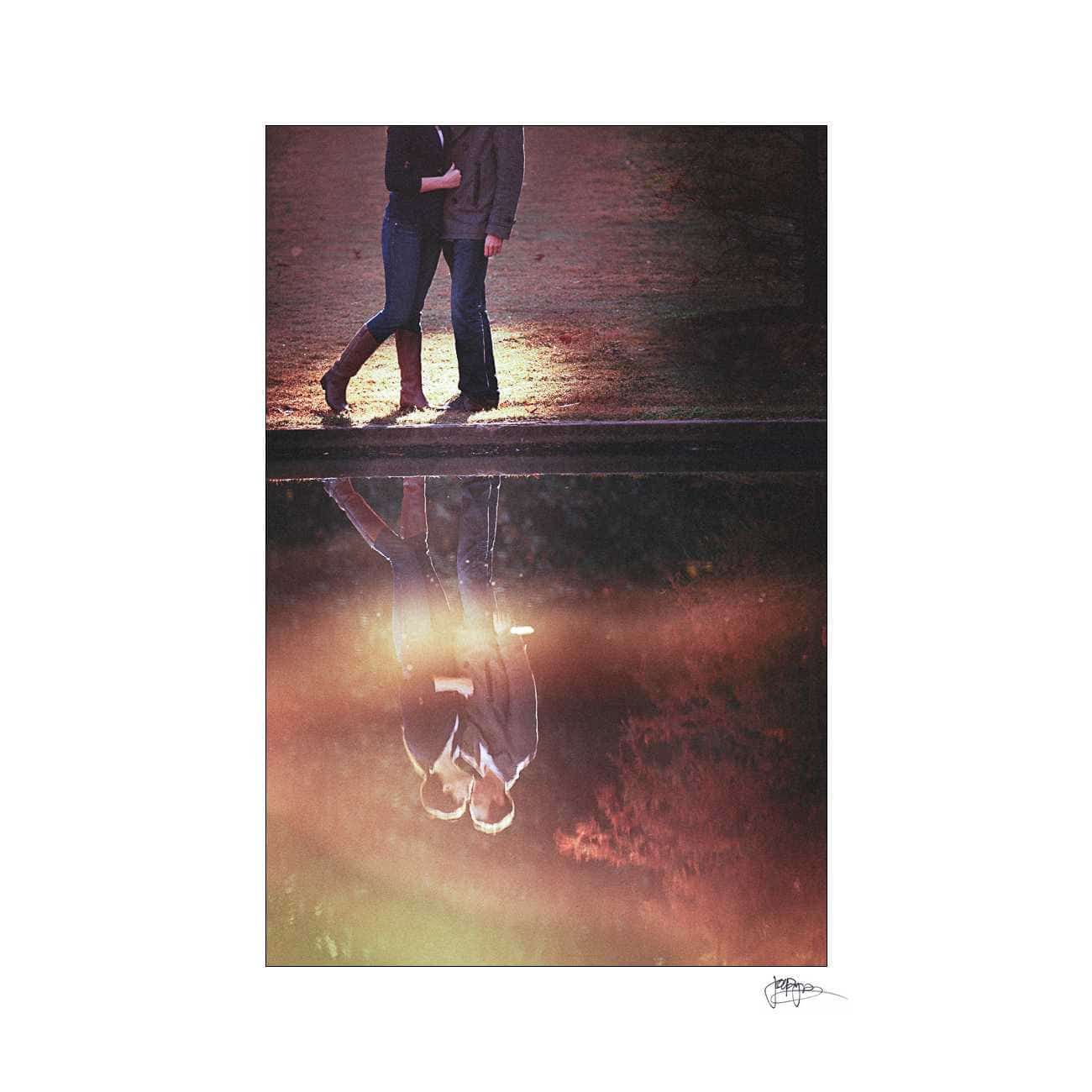 duke chapel engagement photography ~ brianna and chris A couple walks along a path, holding hands, with their reflection visible in a still body of water below. The scene is softly lit with warm, natural tones, creating a serene and romantic atmosphere.