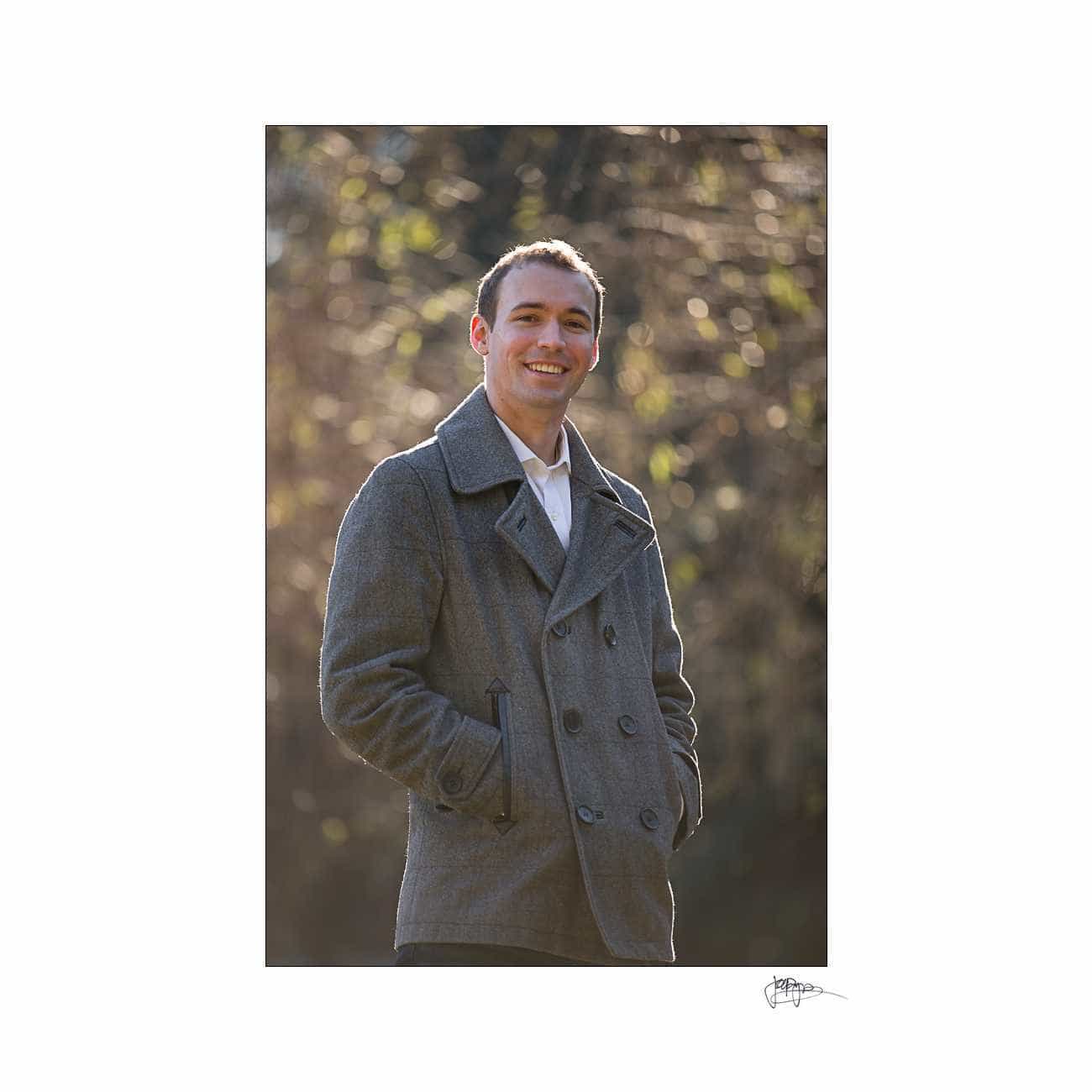 duke chapel engagement photography ~ brianna and chris A man stands outdoors, smiling and wearing a grey coat over a white shirt. The background is softly blurred with hints of greenery and sunlight.