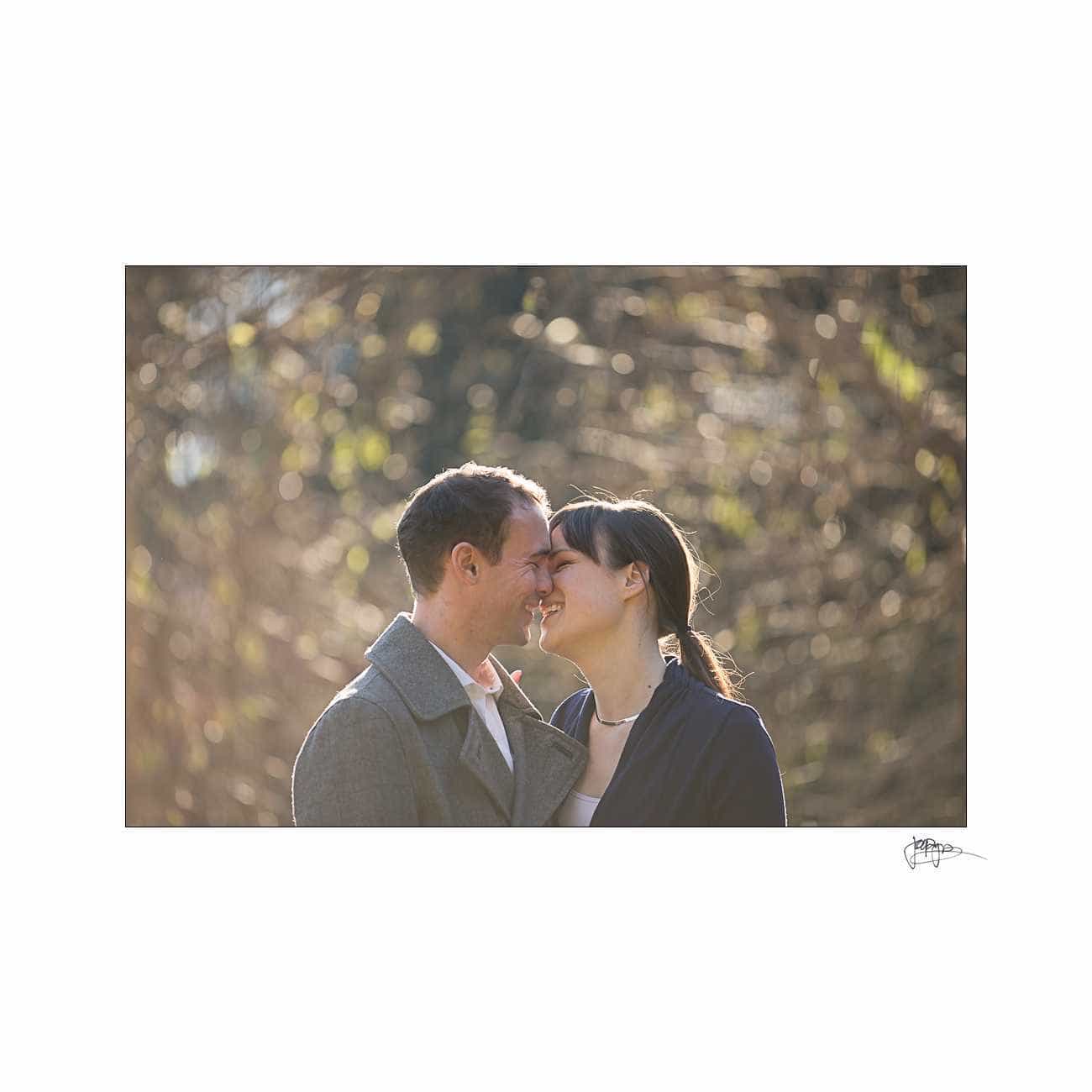 duke chapel engagement photography ~ brianna and chris A couple stands close together, smiling with their foreheads touching. They are outdoors, with blurred trees in the background. The sunlight creates a soft, warm atmosphere. Both are dressed in jackets.