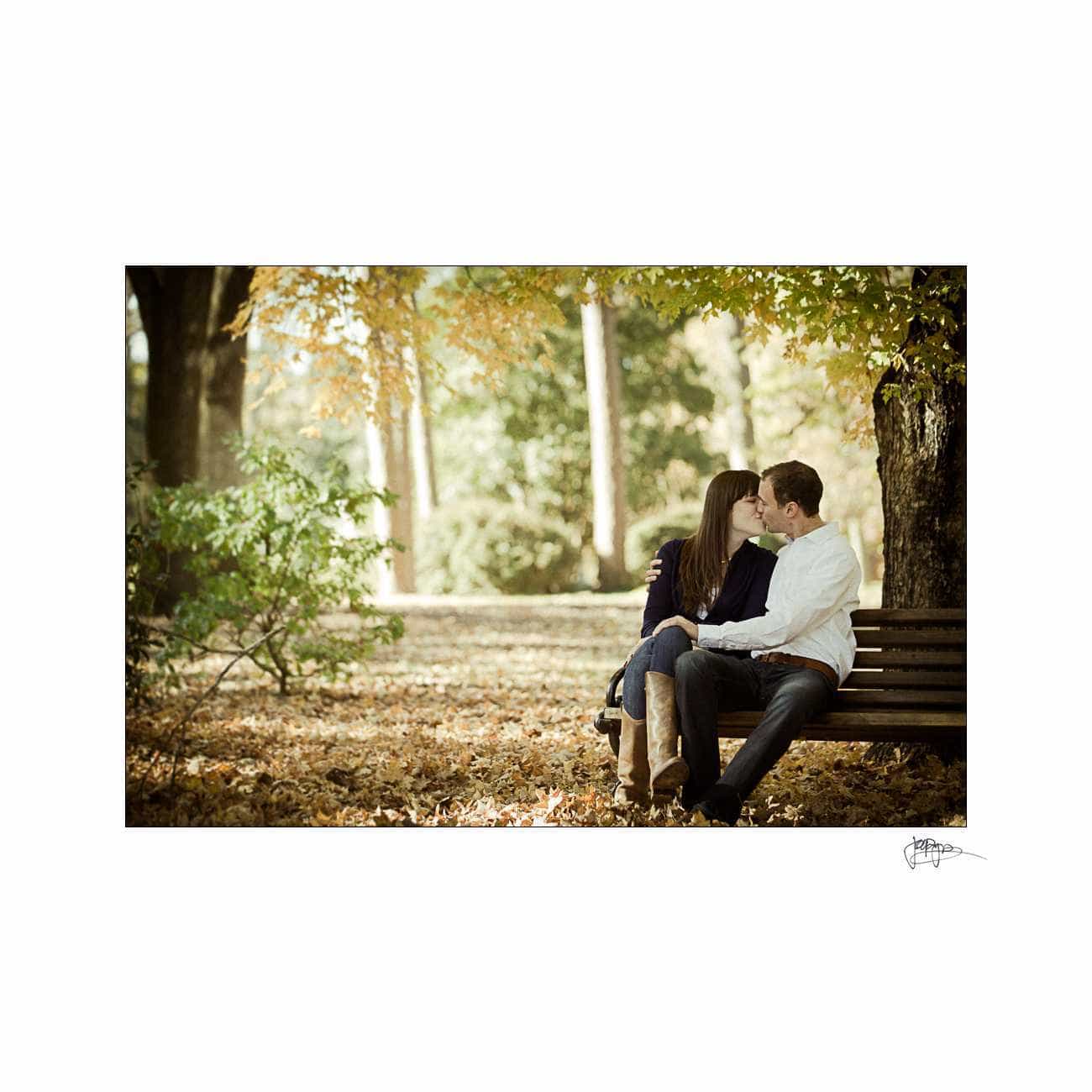 duke chapel engagement photography ~ brianna and chris A couple sits closely on a park bench, surrounded by autumn trees and fallen leaves. They gaze at each other affectionately. The setting is peaceful and sunlit, with green and yellow leaves highlighting the scene.