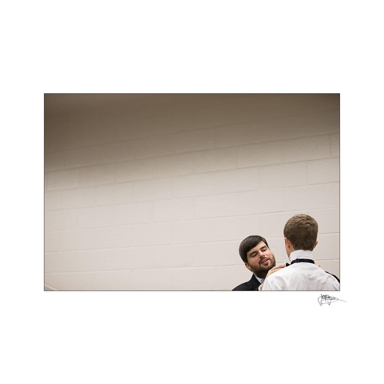 Two men in formal attire are in conversation. One is facing the camera, smiling, while the other has his back turned. The background is a plain, light-colored wall.
