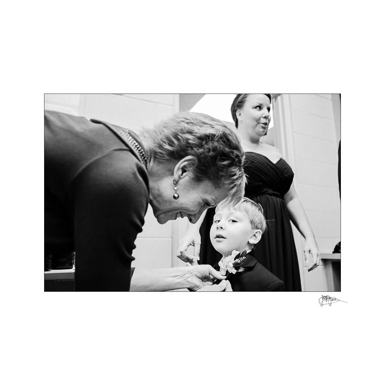 A woman helps adjust a young boys tie or boutonniere while another woman stands in the background, all dressed formally. The scene appears candid and warm, capturing a moment of preparation. The image is in black and white.