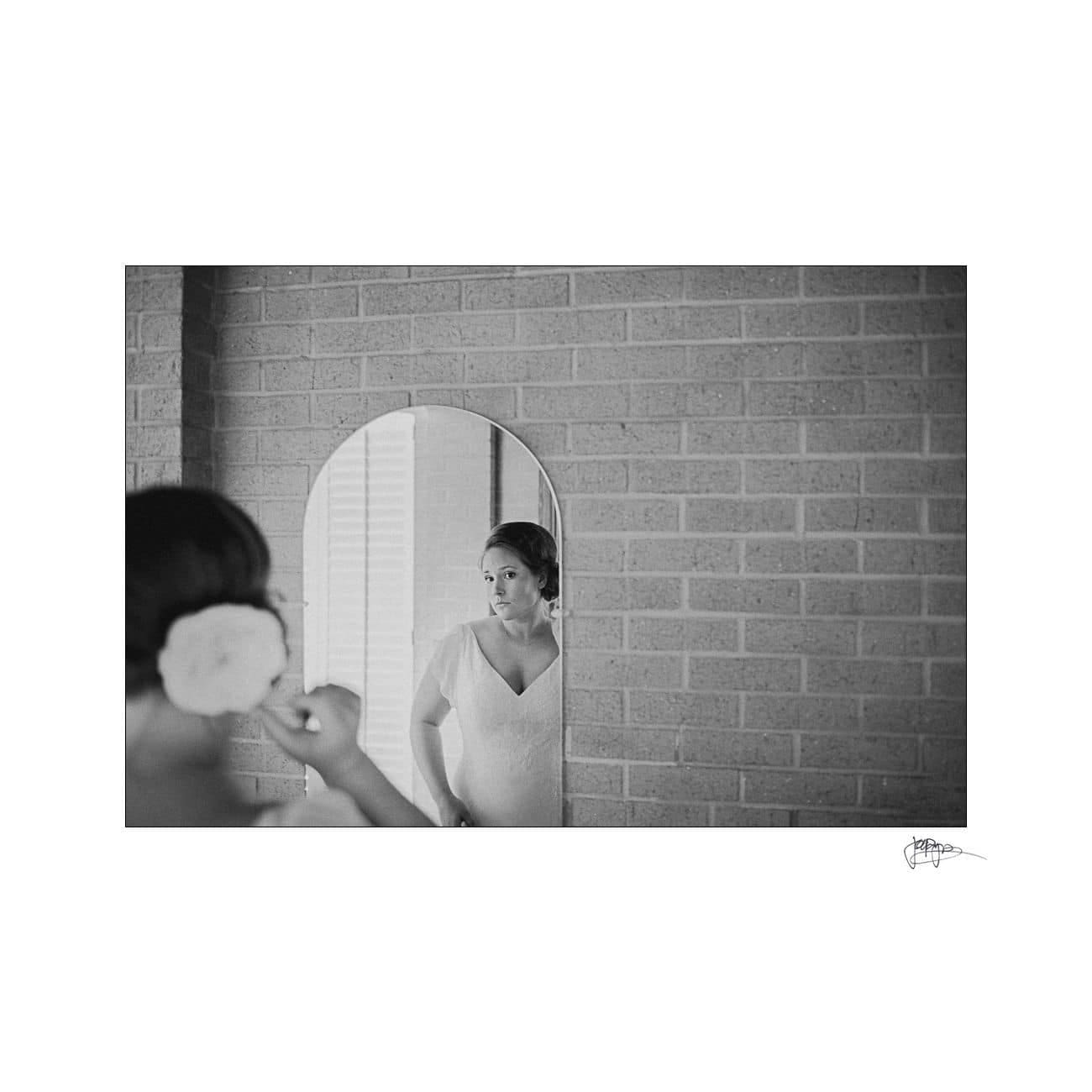 Black and white photo of a bride standing in front of a mirror, adjusting her hair adorned with a flower. Brick wall background and a soft, contemplative expression on her face, partially visible in the mirror.
