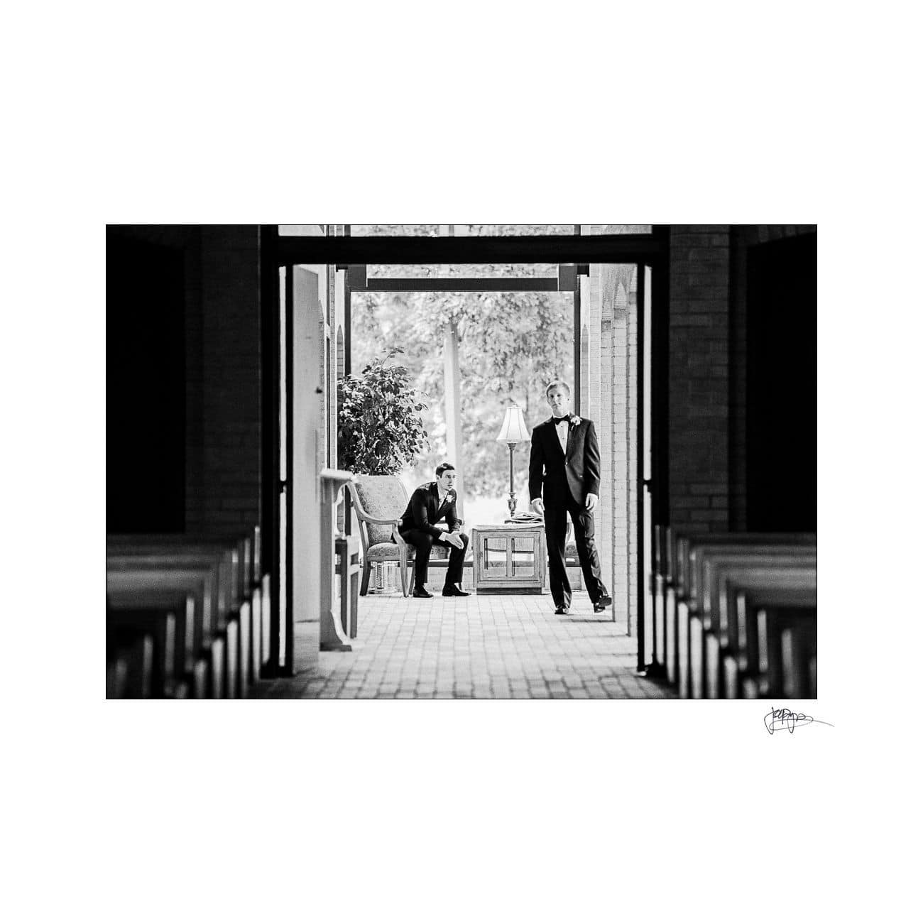 Two men in formal attire are in a corridor with large windows. One is seated in a waiting area, looking down, while the other stands near an open door, gazing outside. The scene is captured in black and white.