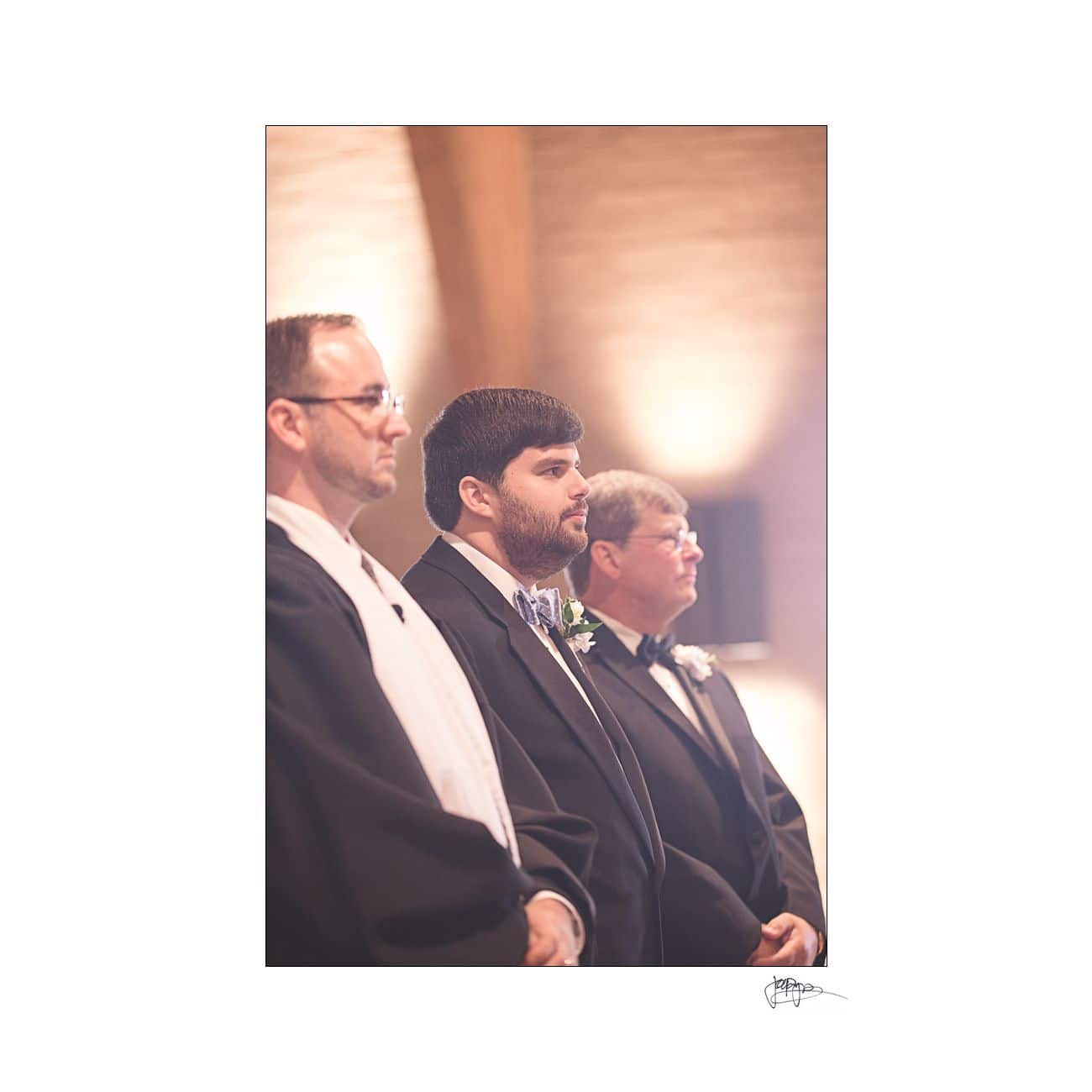Three men in formal attire stand in a row, facing forward. The lighting is warm, highlighting their profiles. The background is softly blurred, emphasizing the men as the focal point of the image.