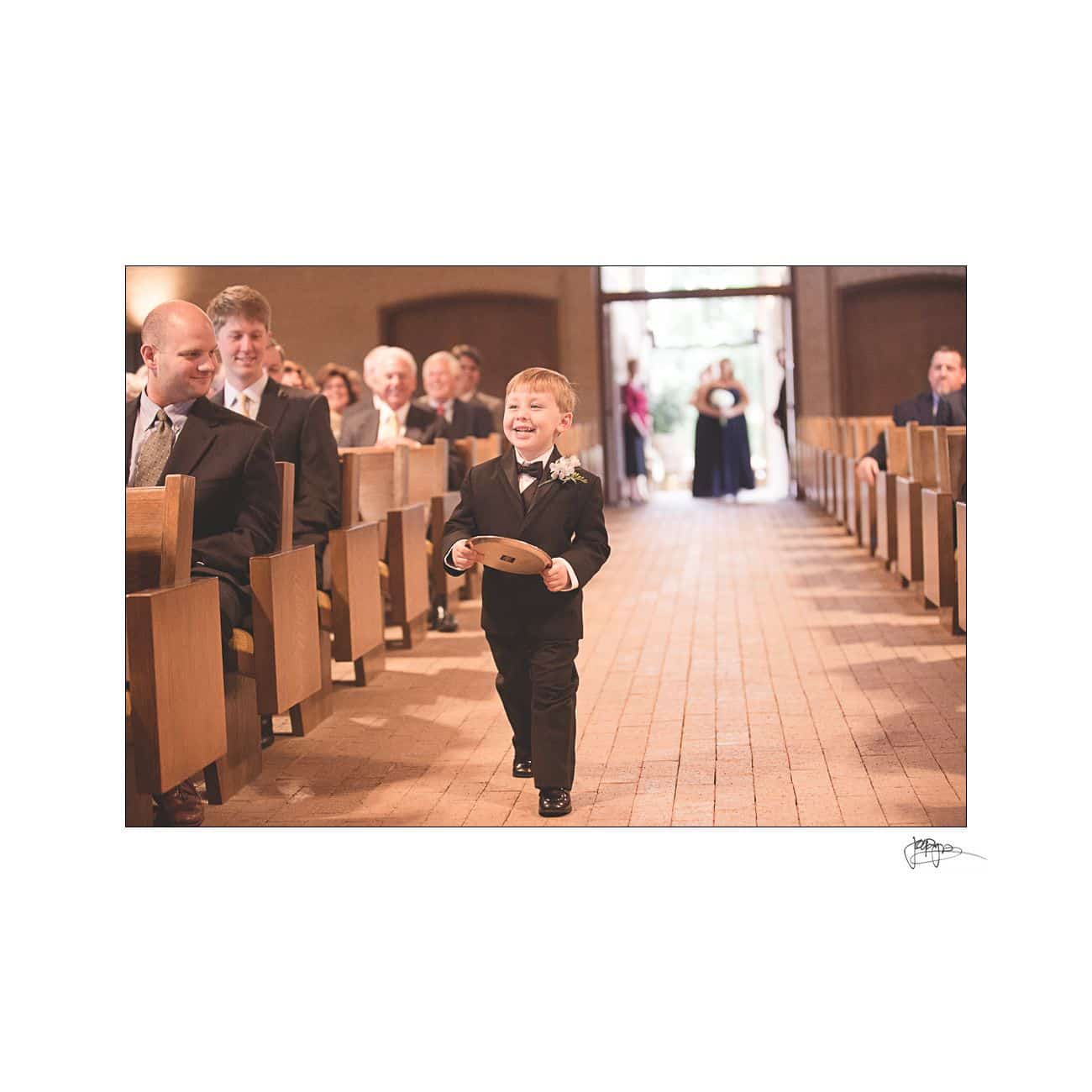 A young boy in a suit walks down a church aisle holding a circular wooden sign with a smile. Guests in formal attire are seated on either side, watching the processional. A person stands at the end of the aisle.