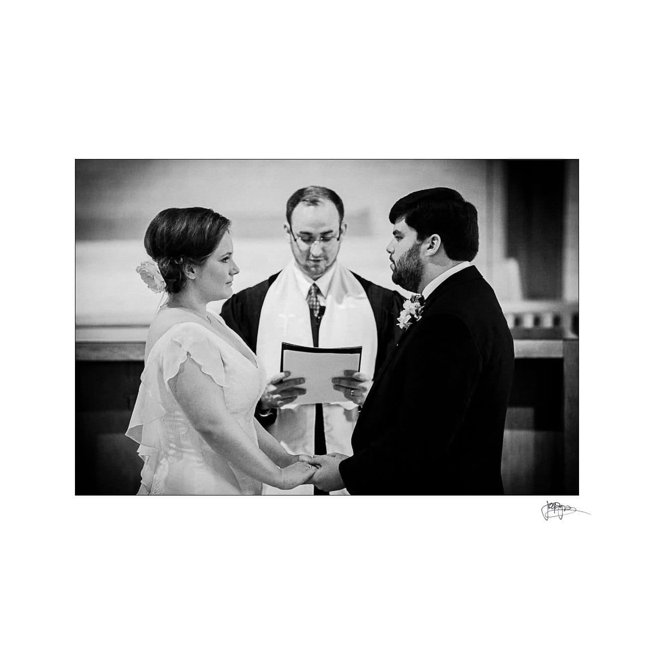 A black and white photo of a bride and groom holding hands, facing each other, with a minister standing in the background reading from a document. The scene appears to be a wedding ceremony in a church setting.