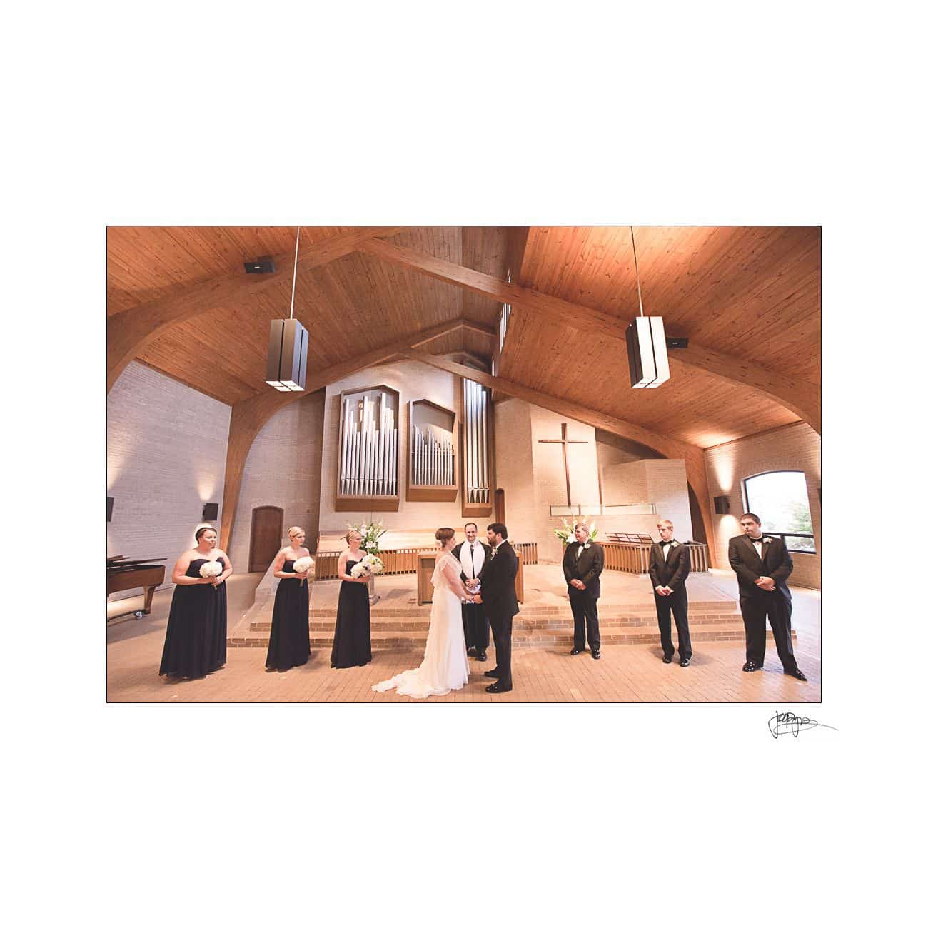 A bride and groom stand at the altar in a church with high wooden ceilings. They are surrounded by bridesmaids in black dresses and groomsmen in black suits, facing the congregation. An organ and a large cross are visible in the background.