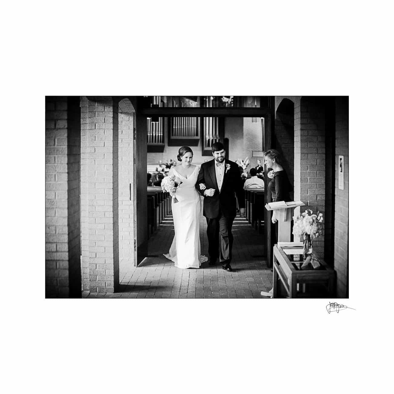 A bride in a long white dress and a groom in a dark suit walk arm in arm down an aisle in a brick church. The setting is elegant, with floral decorations on the side, and seated guests in the background. The image is in black and white.