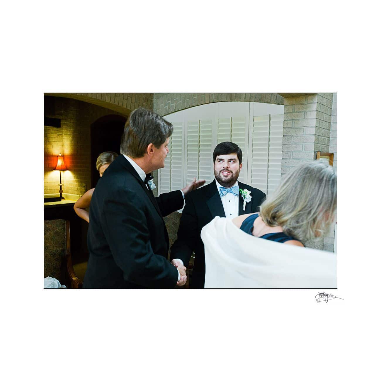 A groom in a tuxedo is greeting a man in a suit with a handshake and a smile. A woman with a white shawl has her back to the camera. Theyre in a warmly lit room with a lamp and shutters in the background.