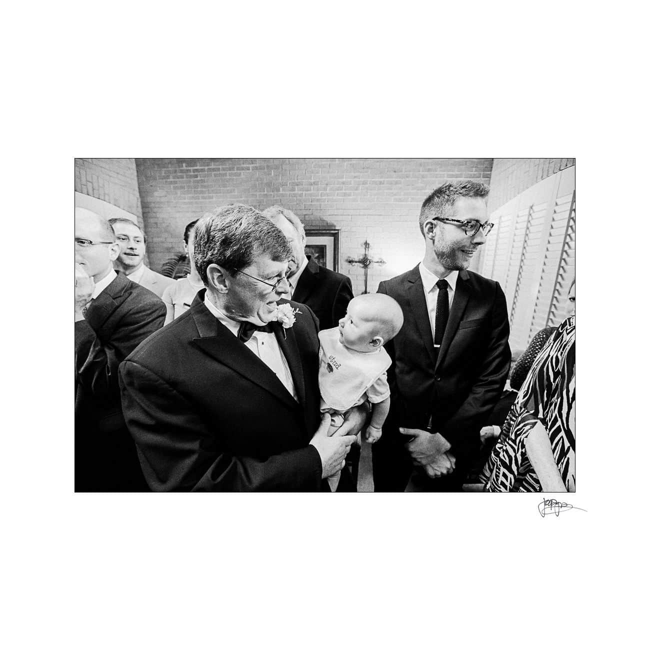 A black and white photo of a formally dressed group at a gathering. An older man in a tuxedo gently holds a baby, surrounded by smiling adults, including a man wearing glasses and a suit, in a warmly lit room.