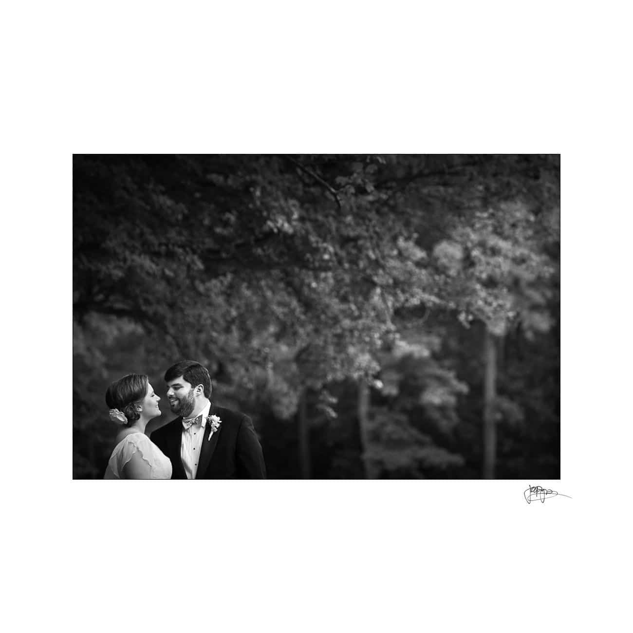 A black and white photo of a couple in wedding attire smiling at each other. They stand outdoors in front of a blurred background of trees. The image has a large white border and a signature in the bottom right corner.