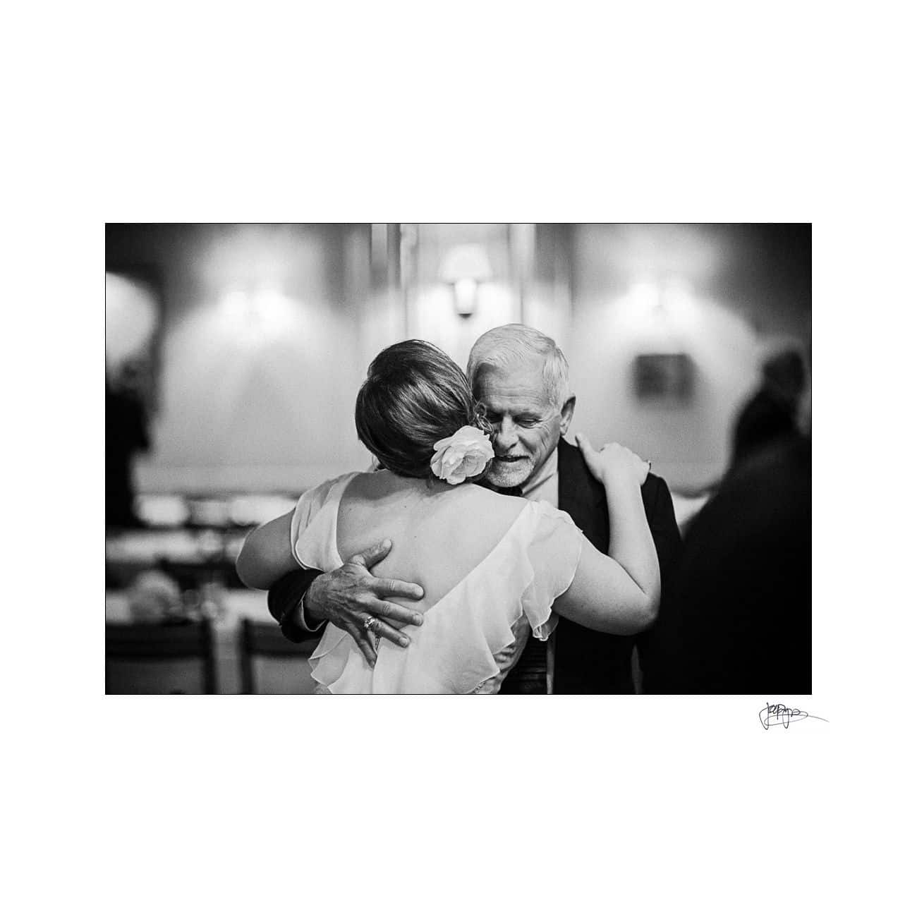 A black and white photo of an older couple dancing closely. The woman, with a flower in her hair, wears a dress with ruffled sleeves. The man is in a suit. They embrace in a softly lit room, exuding a sense of warmth and nostalgia.