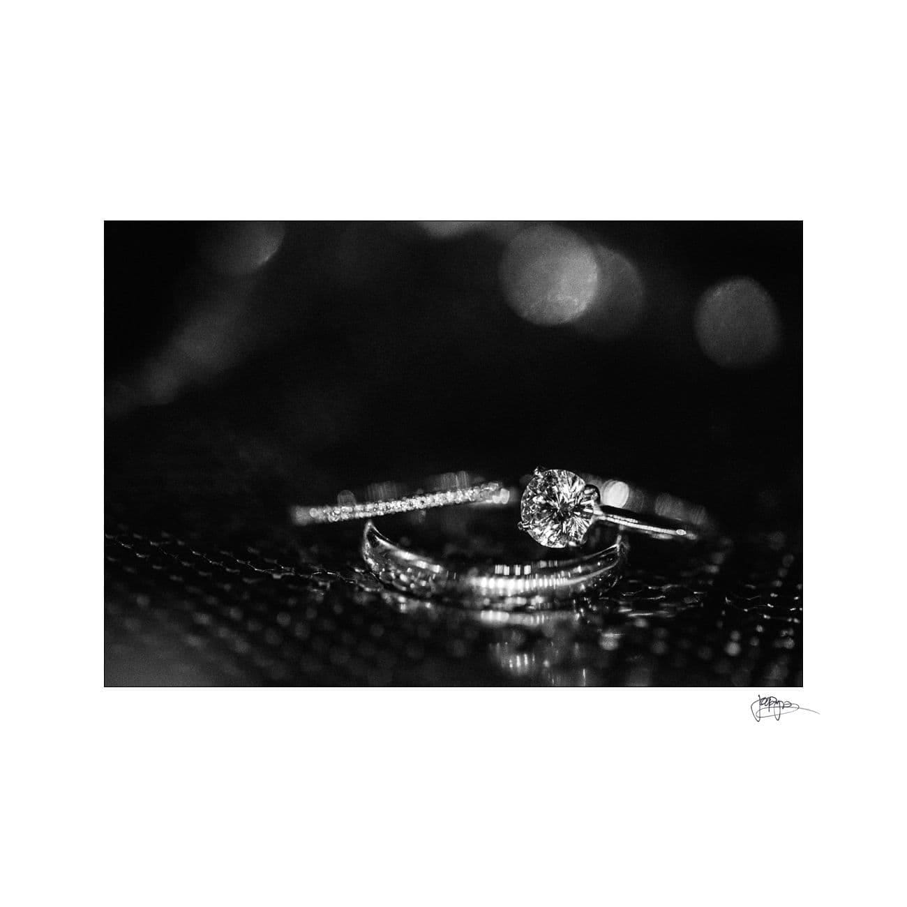 A close-up black and white image of a sparkling diamond engagement ring resting on a textured surface. The ring features a round-cut diamond with several smaller stones adorning the band, capturing light and creating a shimmering effect.