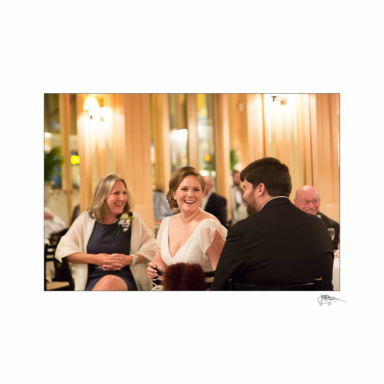 A woman in a white dress smiles and talks with a man in a suit at a dinner event. Another woman with blonde hair sits nearby, also smiling. The setting is warmly lit with elegant decor.