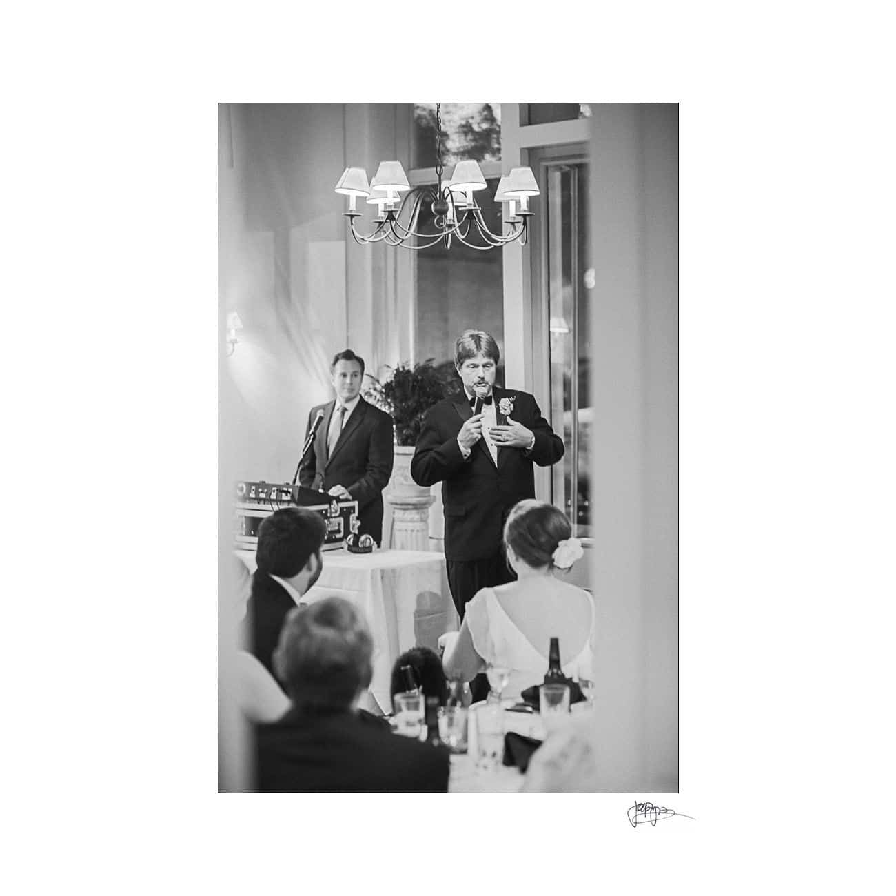 A man in a suit delivers a speech at a formal event, standing near a DJ booth with a microphone. Guests seated at tables listen, and a chandelier hangs from the ceiling. Black and white photo.