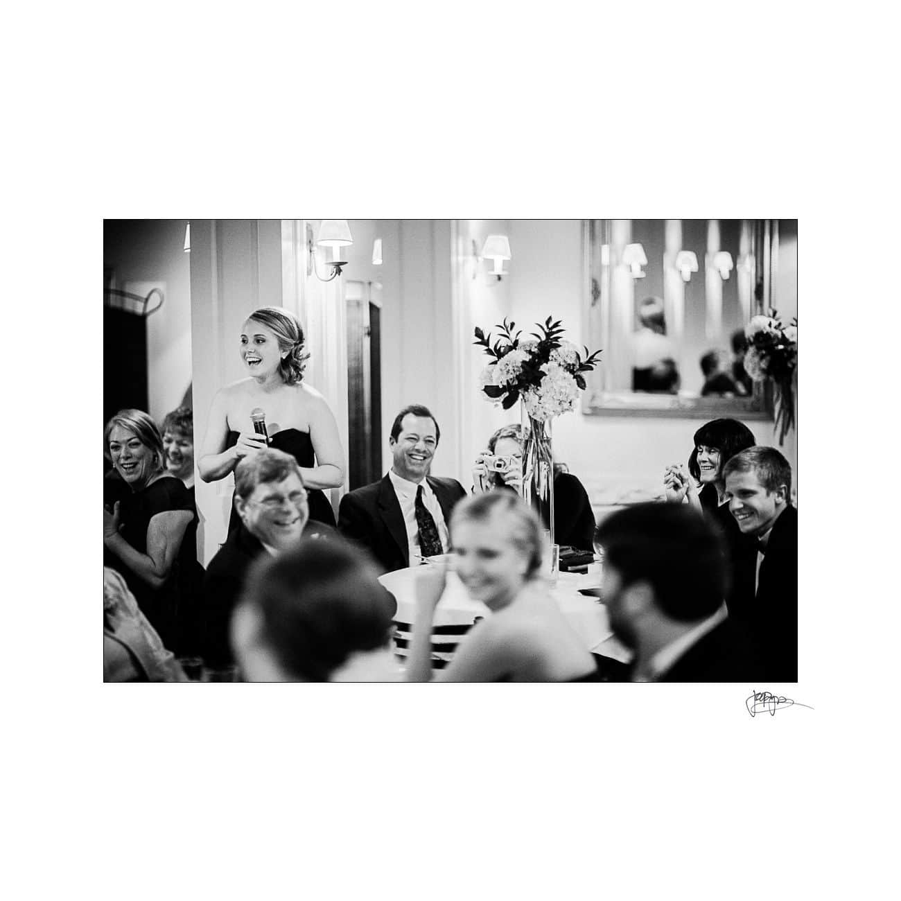 A woman standing and holding a microphone speaks to a seated audience around a table, all smiling, in a formal setting with elegant decor. The image is in black and white.