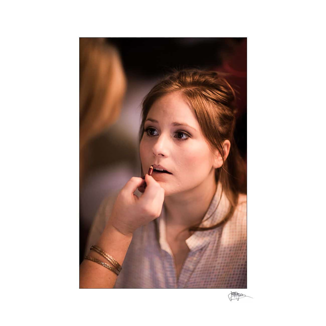 A woman with brown hair is having lipstick applied by another persons hand. She is wearing a light-colored top and looking slightly upward, with a focused expression. The background is softly blurred.