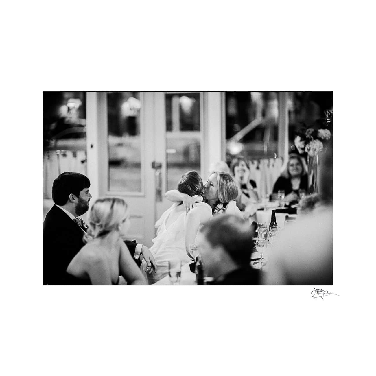 A black and white photo of a wedding reception. In the foreground, a couple is seated at a table. In the background, a bride is hugging a guest, with more guests seated and chatting nearby. There are flowers and drinks on the tables.