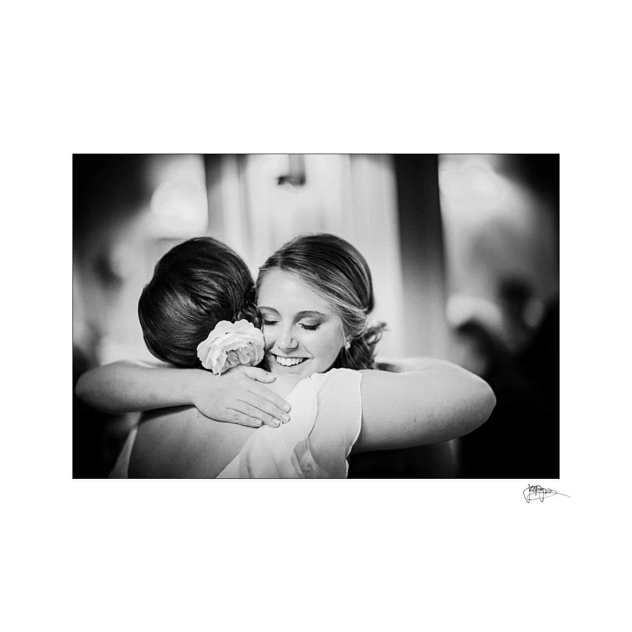 A black and white photo of two women embracing warmly. One woman has a flower in her hair, and both appear happy and content. The background is softly blurred, highlighting their affectionate moment.