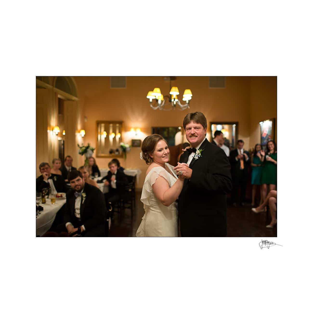 A bride and groom dance together in a warmly lit room. Guests seated and standing in the background watch and smile. The room is decorated elegantly with flowers and soft lighting.