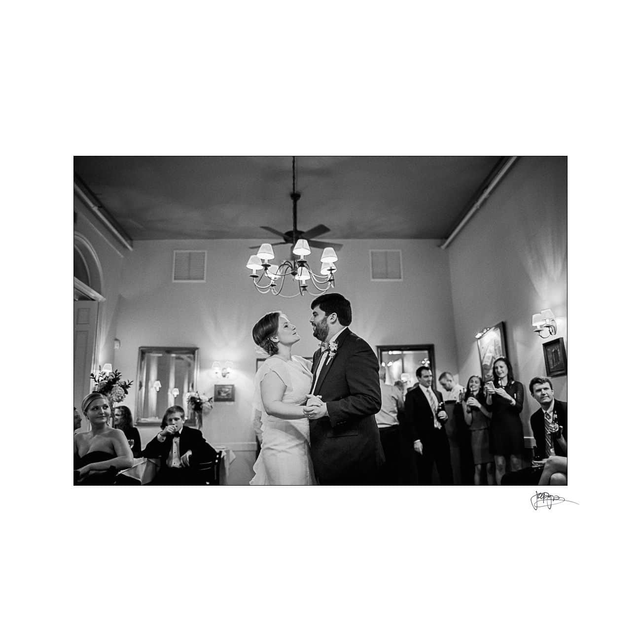 A black-and-white photo of a couple sharing a dance in a formal setting, surrounded by smiling guests. The bride wears a wedding dress and the groom a suit. A chandelier hangs above them, creating a warm ambiance.