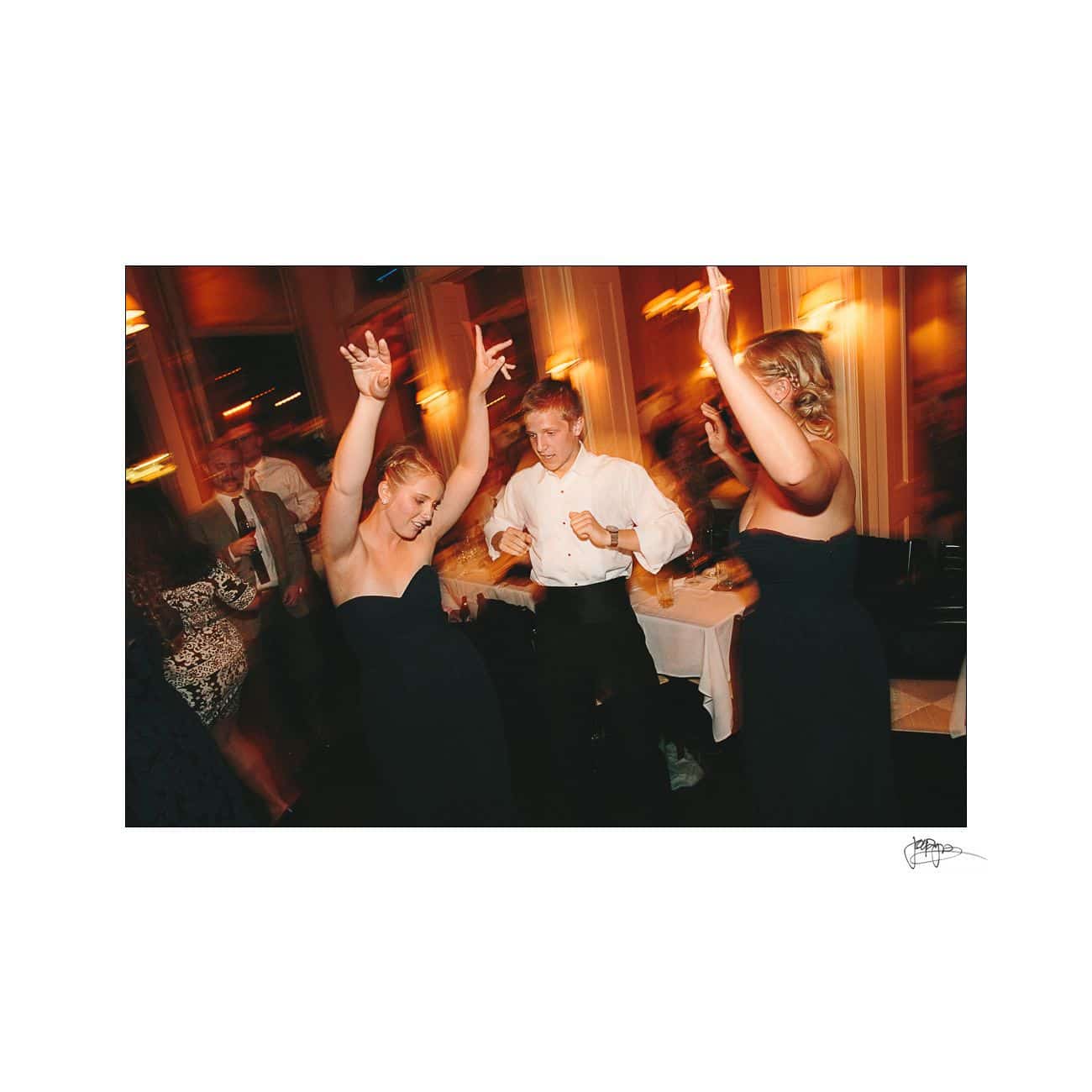 People are dancing joyfully at an indoor event. Two women in black dresses and a man in a white shirt are in the foreground with their arms raised. The background is softly blurred with warm lighting, creating a festive atmosphere.
