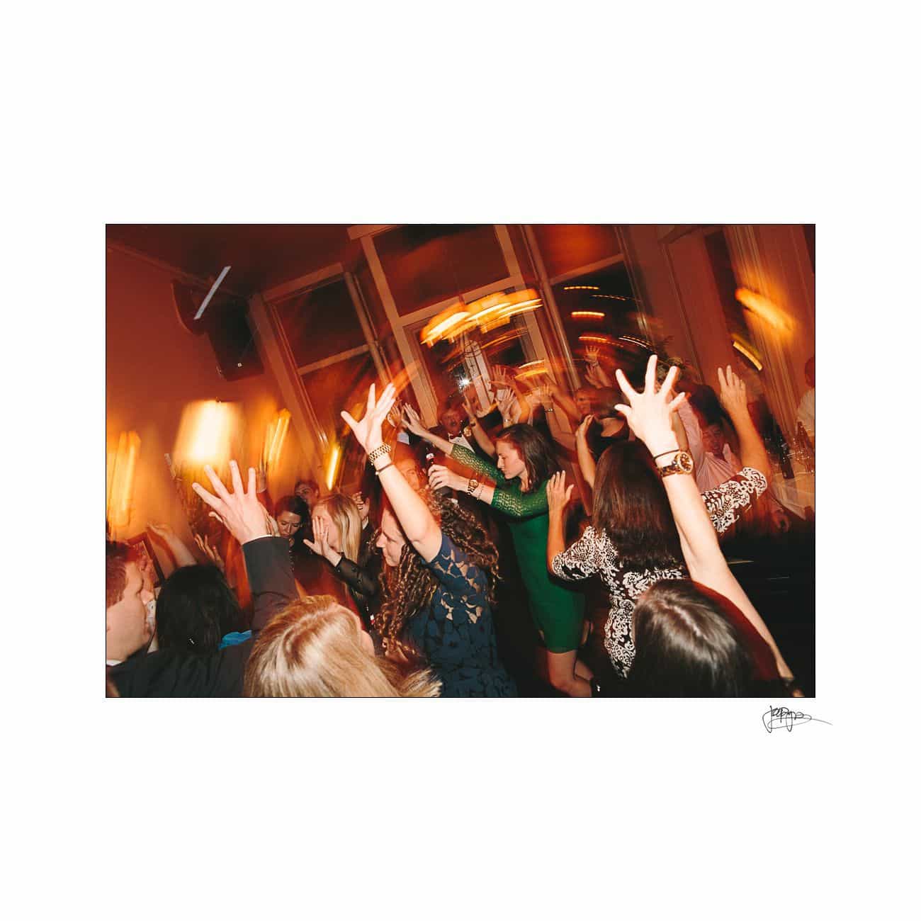 A lively group of people with arms raised are dancing energetically in a warmly lit room. Blurred motion suggests movement and joyful activity at a party or celebration.