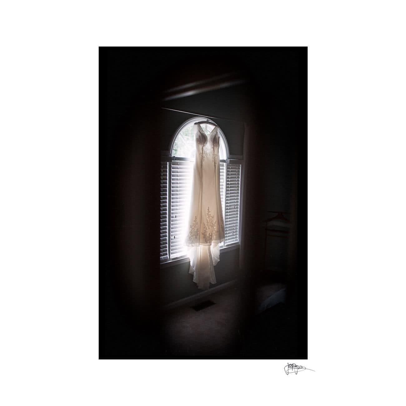 A white wedding dress hangs elegantly in front of a tall, arched window with closed blinds. Soft light filters through, highlighting the delicate details of the dress. The room is dimly lit, creating a serene and intimate atmosphere.